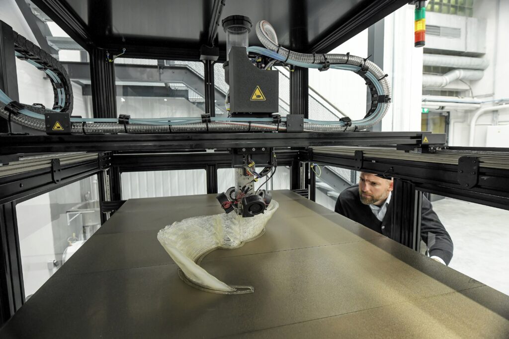 Didelis 3D spausdintuvas kuria skaidrų banguojantį objektą, vyras stebi.