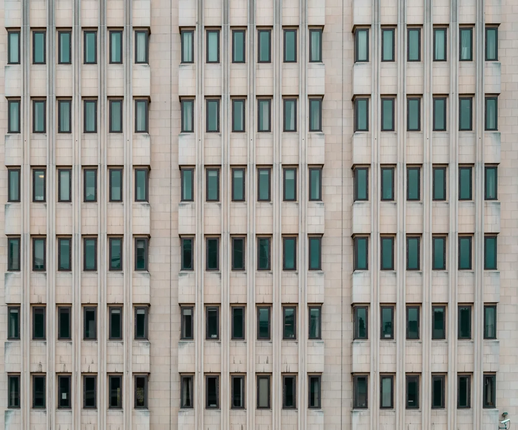 Vienodos eilės tamsių langų blyškios smėlio spalvos fasade pabrėžia biuro pastato monotoniją ir simetriją.