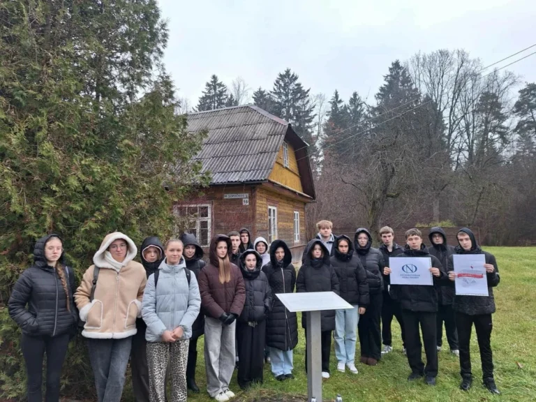 Paauglių grupė šiltai apsirengę pozuoja prie seno medinio namo, lauke tvyro apsiniaukęs rudens oras.