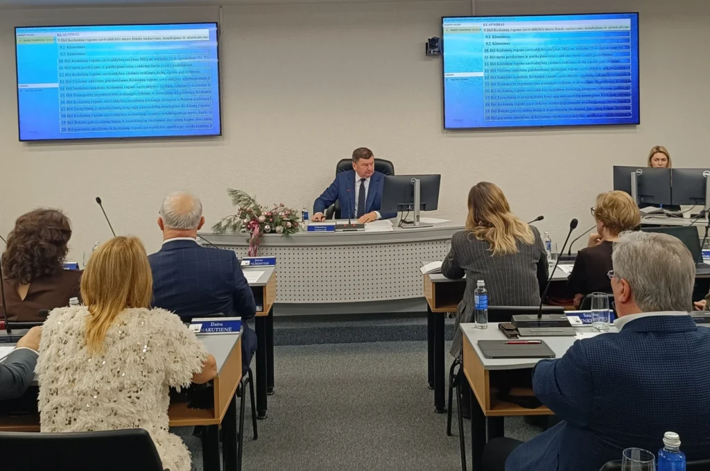 Formalus tarybos posėdis: vyras šviesiai mėlyname kostiume kalba, aplink jį susirinkę nariai, virš galvos šviečia darbotvarkė.