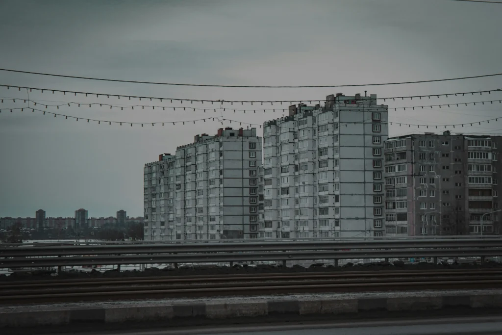 Niūri miesto panorama su pilkais sovietiniais daugiabučiais, kontrastuojančiais su virš tramvajaus bėgių pakabintomis girliandomis.