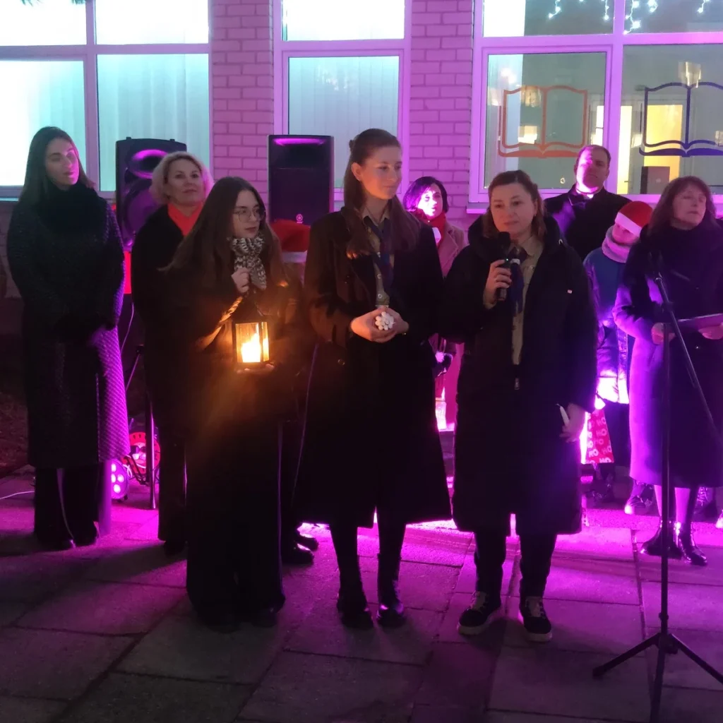 Violetinėmis scenos šviesomis apšviesta moterų grupė mini renginį; viena laiko šviečiantį žibintą.