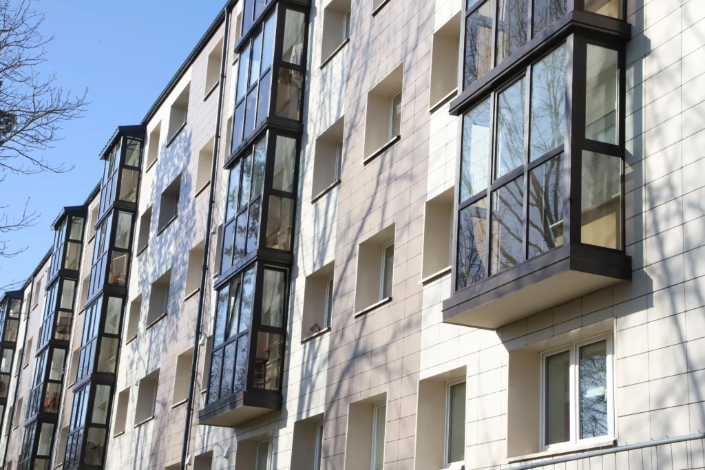 Šviesus modernaus daugiabučio fasadas su stiklintais balkonais ir ryškiais medžių šakų šešėliais.