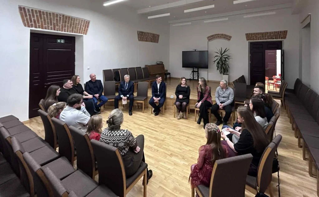 Erdvioje, šviesioje salėje dvidešimt žmonių sėdi dideliu puslankiu ant rudų kėdžių, įsitraukę į aktyvią diskusiją.