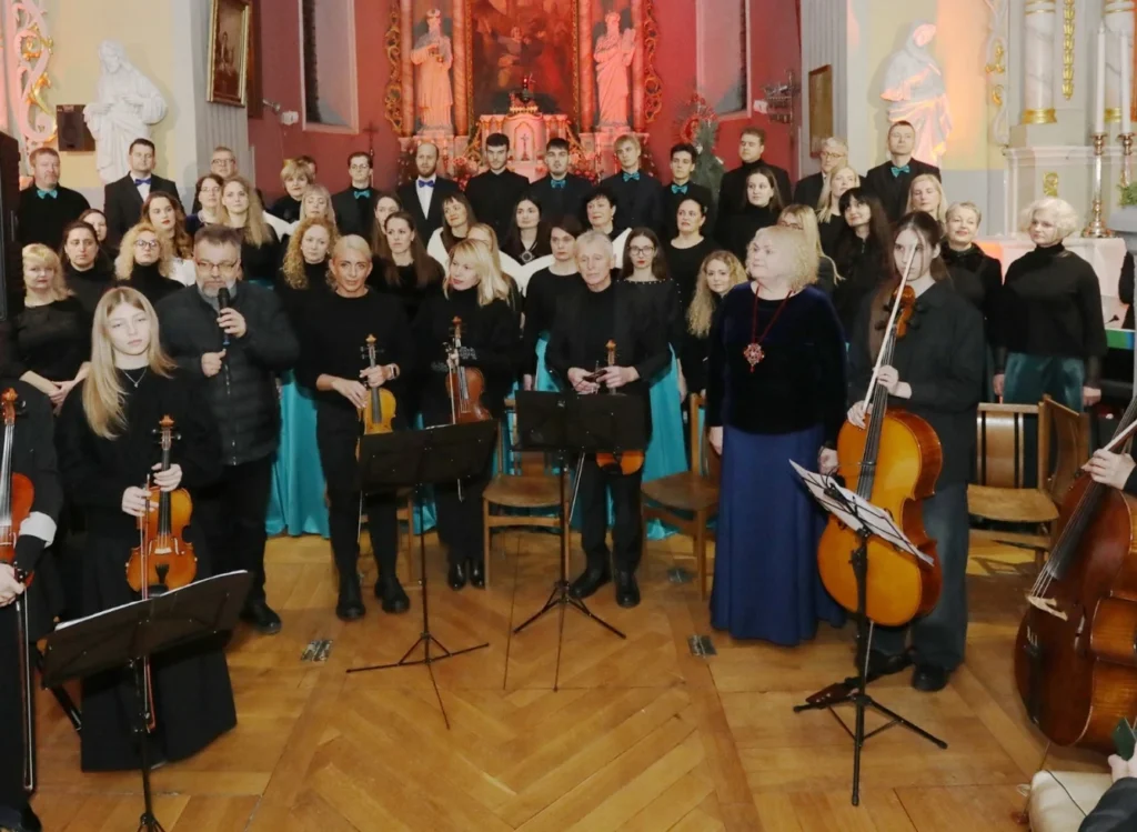 Didelis choras ir styginių instrumentų atlikėjai juodais drabužiais koncertuoja senosios bažnyčios erdvėje su raudonu altoriumi fone.
