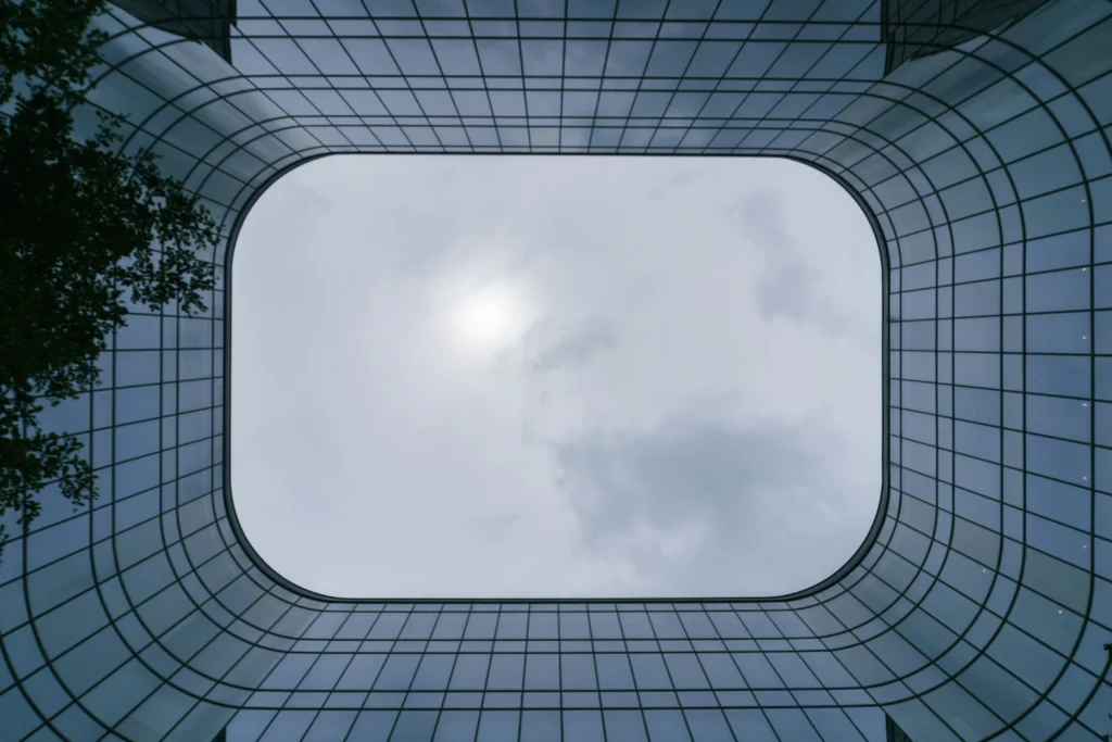 Iš apačios nufotografuotos modernaus pastato išlenktos stiklo sienos įrėmina apšviestą debesuotą dangų.