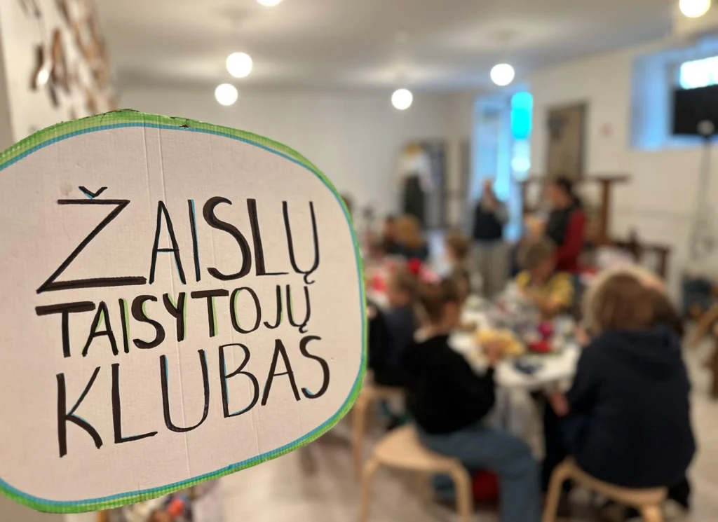 Ryškus kartoninis užrašas „Žaislų Taisytojų Klubas“ priekyje, fone vaikai taiso žaislus bendruomenės erdvėje.