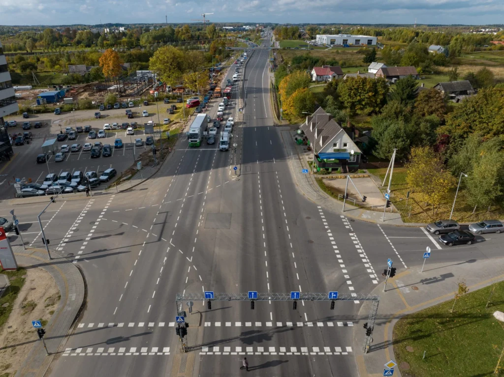 Aukštumos kadras užfiksuoja rudeninę keturių juostų sankryžą, kurioje driekiasi automobilių ir sunkiasvorių vilkikų spūstis, apsupta statybviečių ir žaliuojančių medžių.