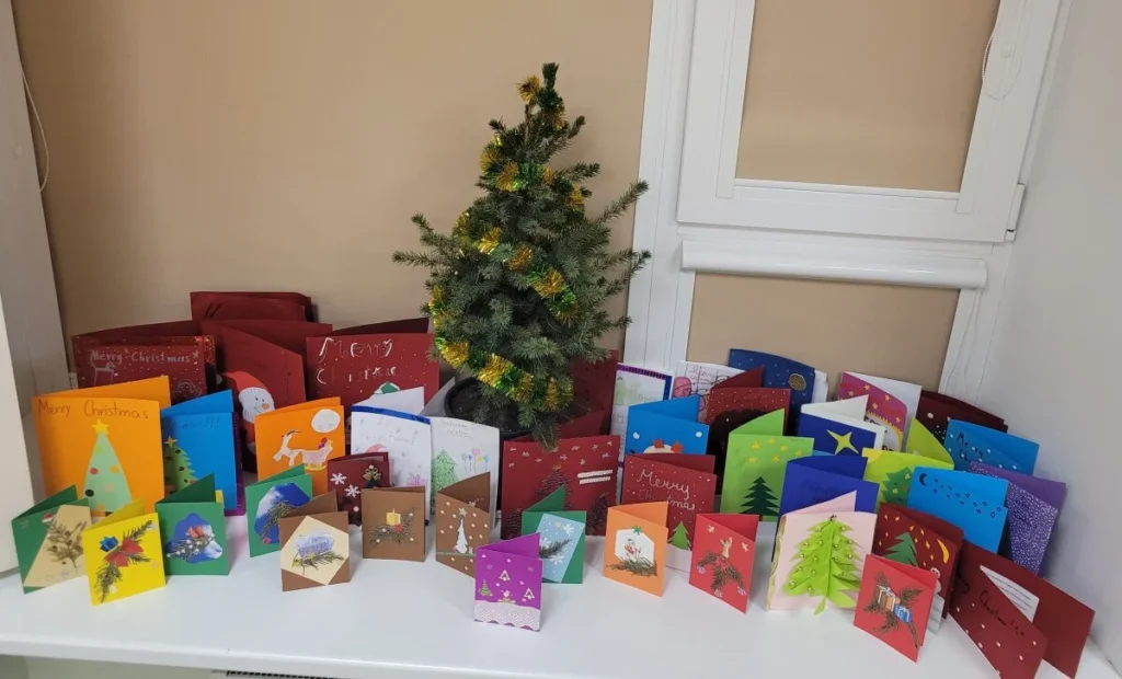 Ryškiaspalviai rankų darbo kalėdiniai atvirukai supa blizgančia girlianda papuoštą eglutę ant balto stalo.