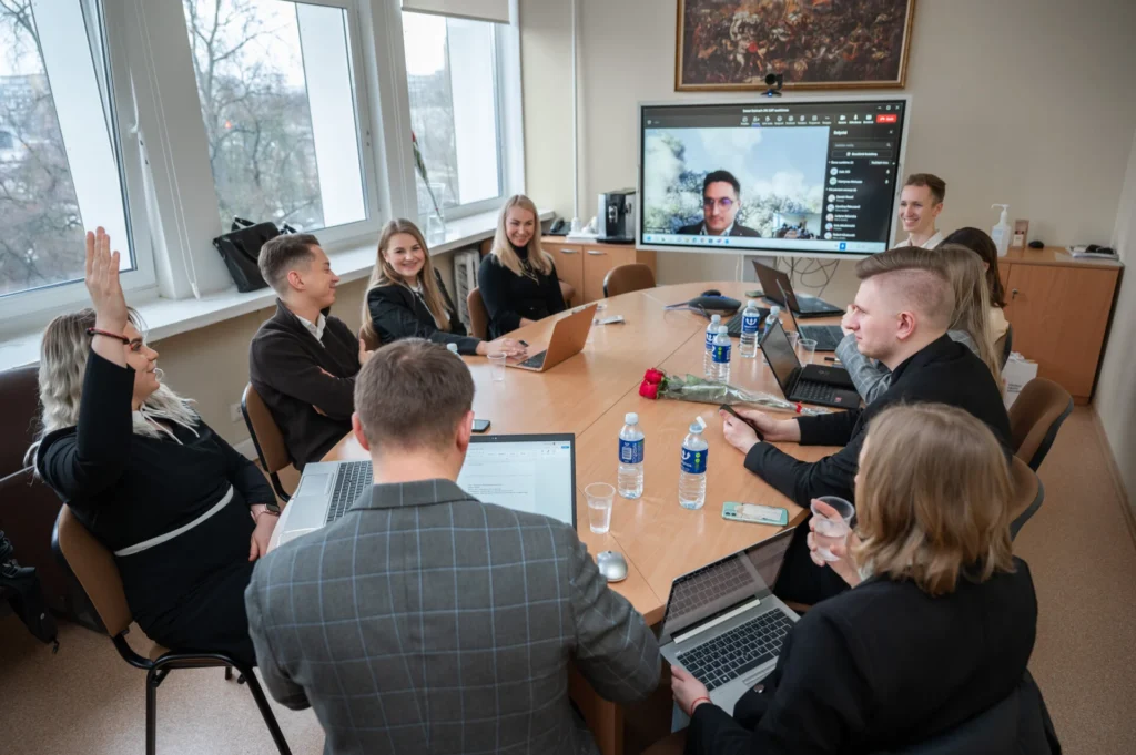 Darbo susitikimas: grupė jaunų specialistų prie stalo, vienai keliančiai ranką, ir nuotolinis dalyvis ekrane.