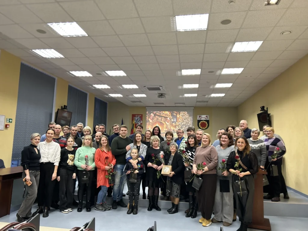 Apie penkiasdešimt žmonių, įskaitant moteris su rožėmis, pozuoja oficialioje salėje su šventine projekcija fone.