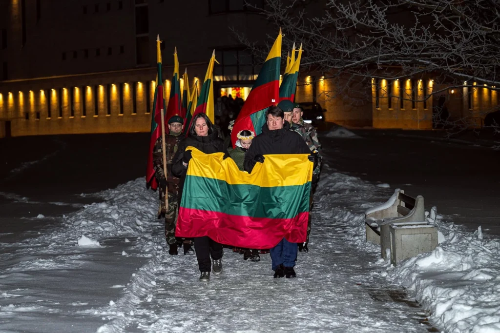 Tamsią žiemos naktį, snieguota alėja pora neša didžiulę Lietuvos trispalvę, lydima vėliavnešių.