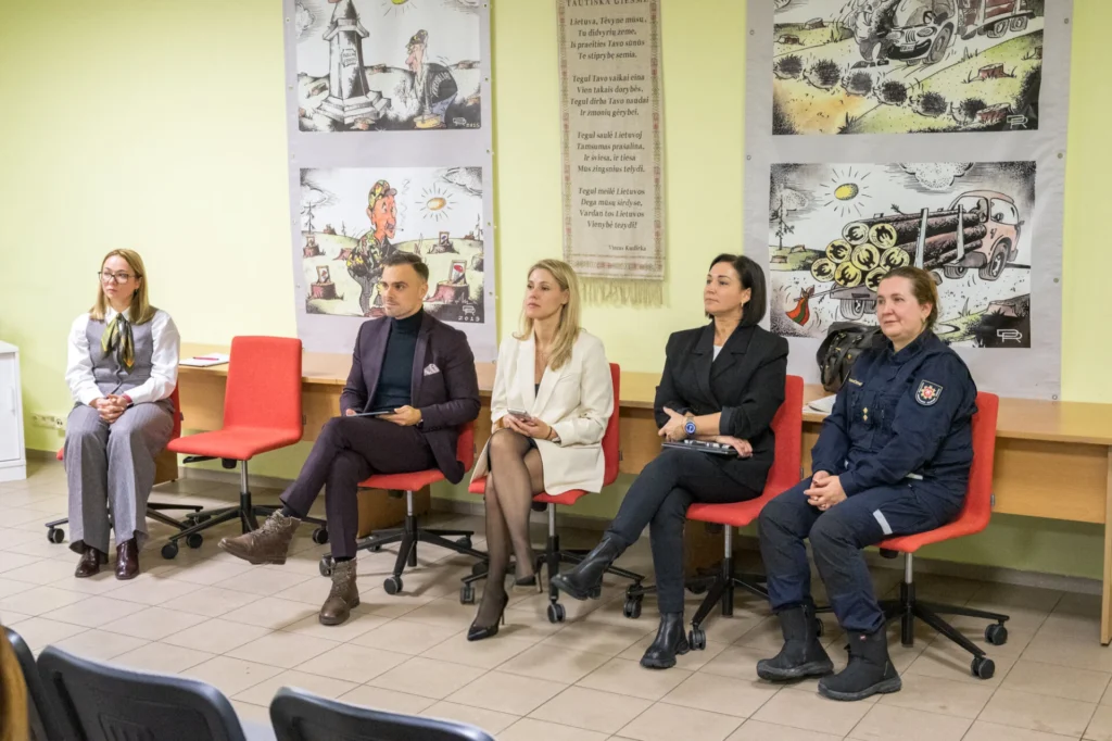 Panelė: penki asmenys, įskaitant pareigūnę, sėdi po Lietuvos himno ir kritiškų miško kirtimo karikatūrų plakatais.