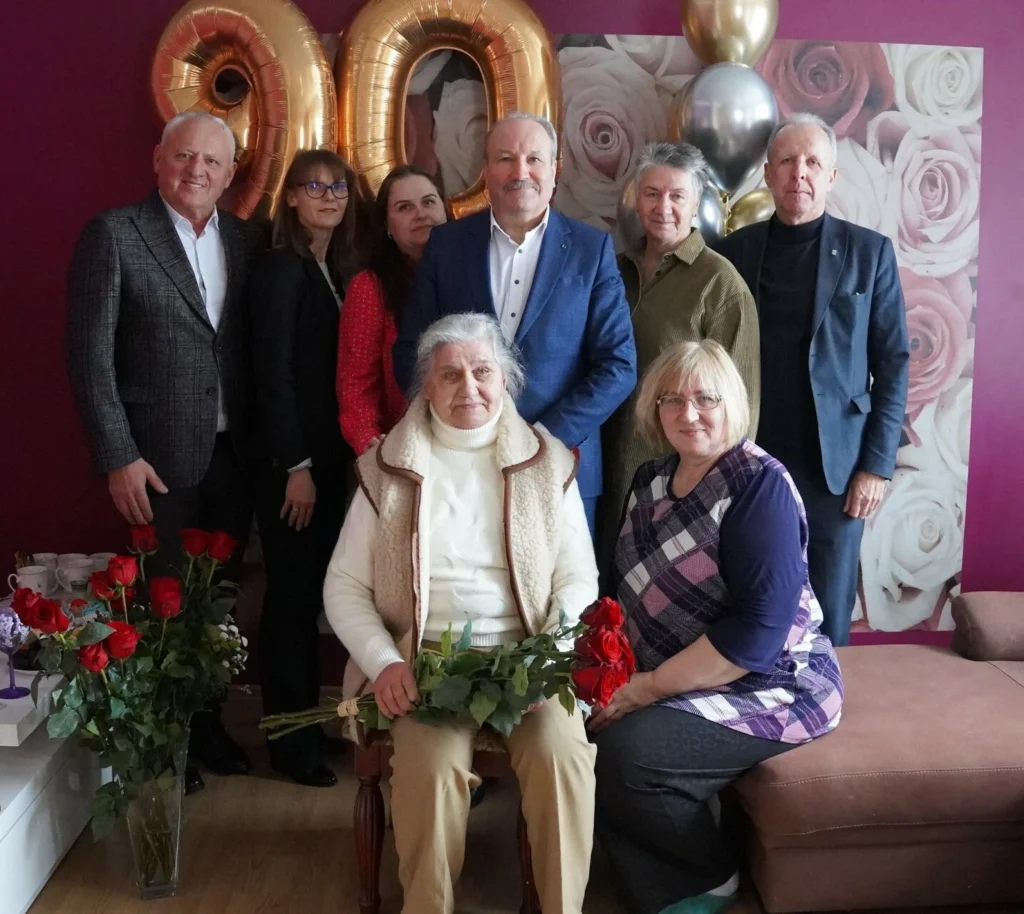 Šeimos nuotrauka: sėdinti garbi moteris, švenčianti 90-metį, apsupta artimųjų, rožių ir auksinių balionų.