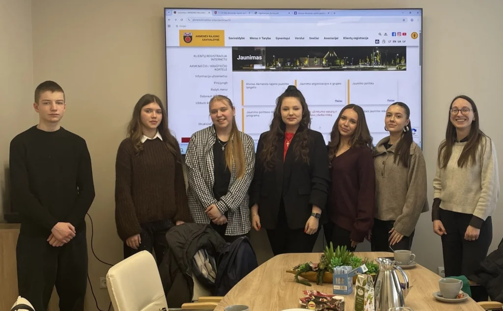 Septyni besišypsantys jaunuoliai stovi priešais ekraną, kuriame rodomas Akmenės rajono savivaldybės jaunimo informacinis puslapis.
