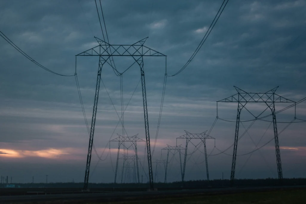 Aukštos įtampos elektros perdavimo linijų stulpai, besidriekiantys į tolį temstant po debesuotu dangumi.