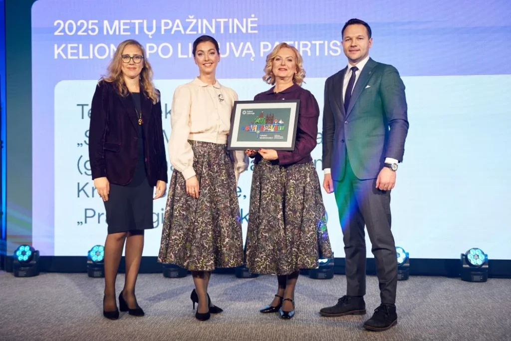 Trys moterys ir kostiumuotas vyras scenoje pristato įrėmintą 2025 m. kelionių po Lietuvą vizualą, šviesiame fone.