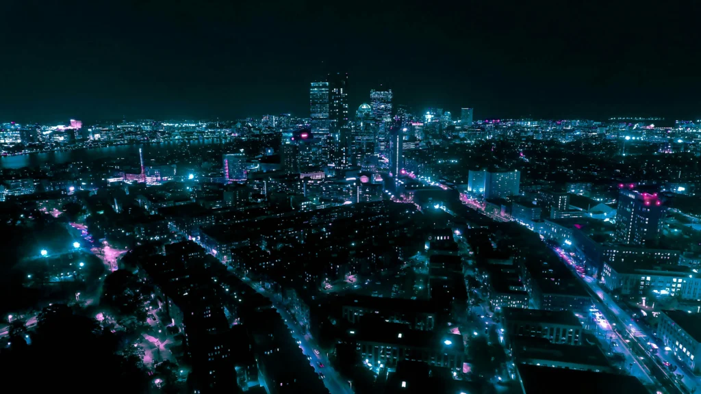 Naktinė miesto panorama iš viršaus su apšviestais dangoraižiais ir ryškiomis neoninėmis melsvos bei violetinės spalvų šviesomis.