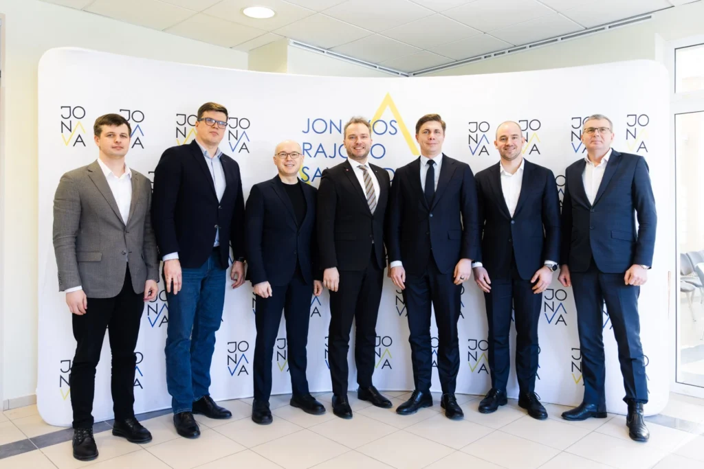 Oficialus septynių vyrų, vilkinčių tamsius kostiumus, portretas prie „Jonavos rajono savivaldybės“ reklaminio stendo.