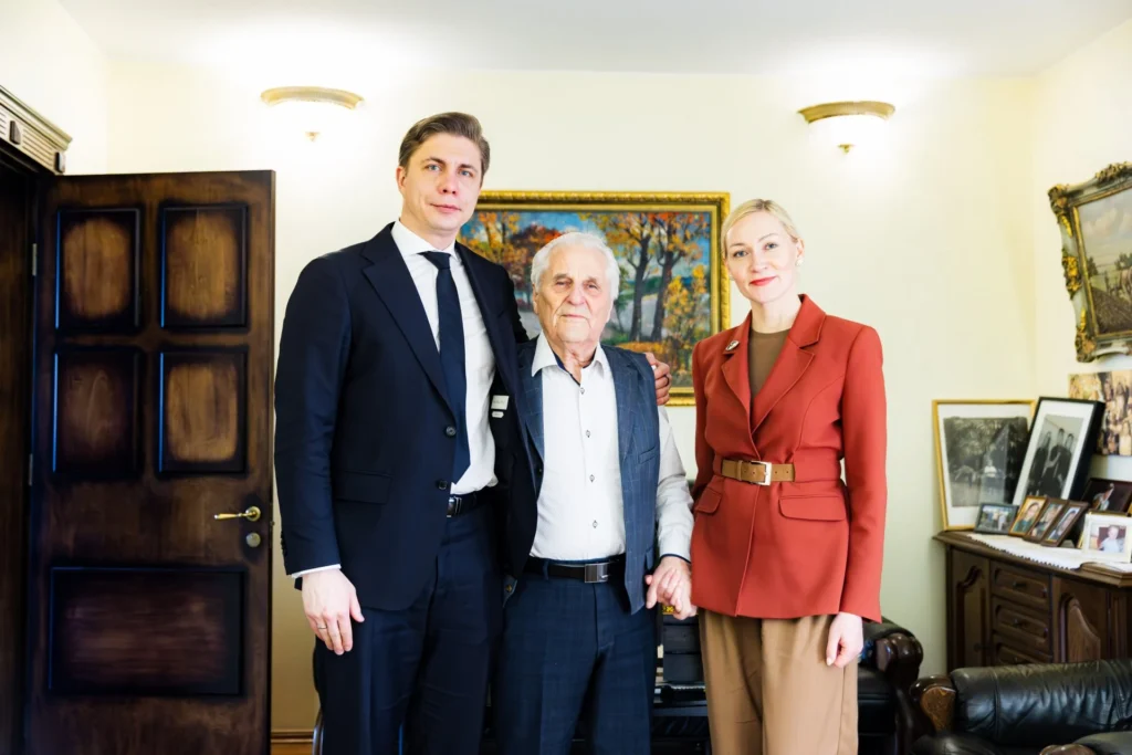 Elegantiškai apsirengę prezidentinė pora, apsikabinusi senolį, pozuoja šiltoje klasikinėje aplinkoje.