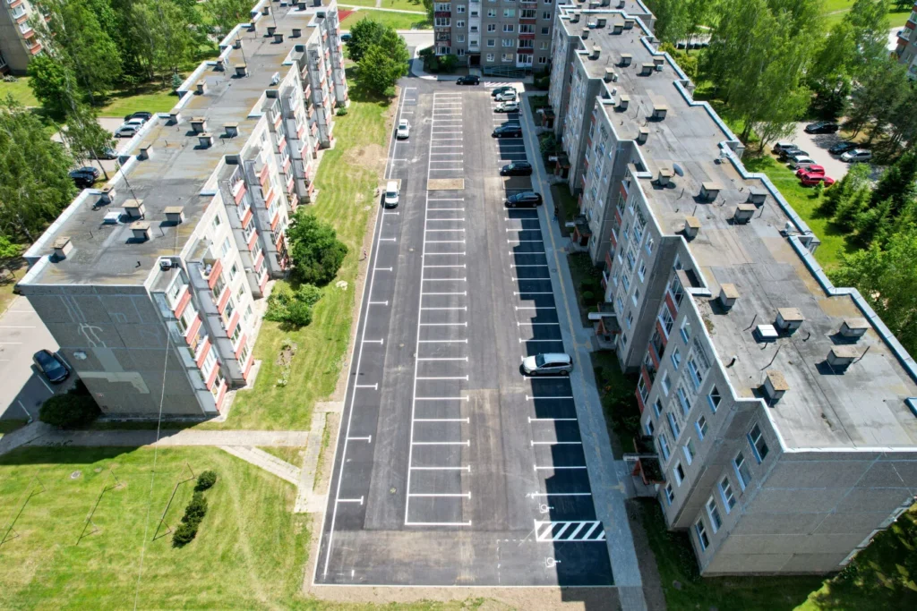 Naujai išasfaltuota ilga automobilių stovėjimo aikštelė tarp dviejų tarybinių daugiabučių, nufotografuota iš viršaus.