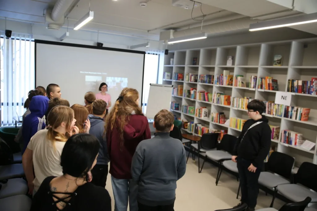 Būrys jaunuolių bibliotekoje žiūri į projektoriaus ekraną, šalia pilnos knygų lentynos ir vadovė.