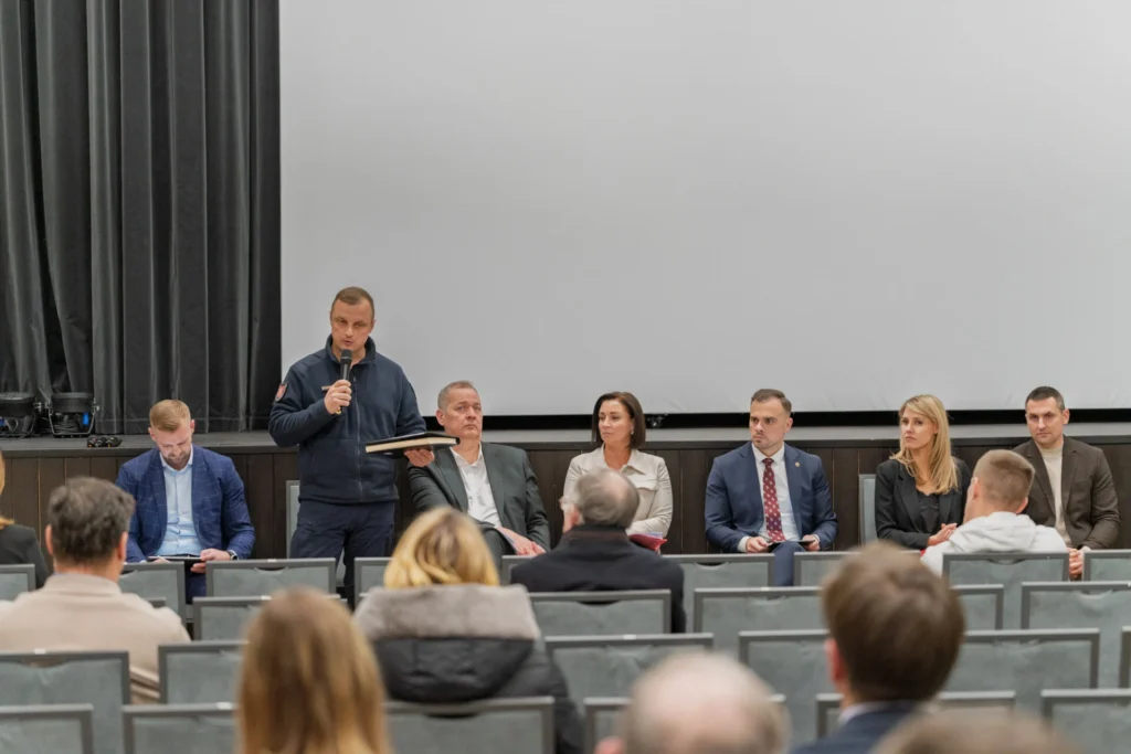 Uniformuotas pareigūnas su mikrofonu kalba auditorijai, už jo sėdi oficialiai apsirengusių asmenų grupė.
