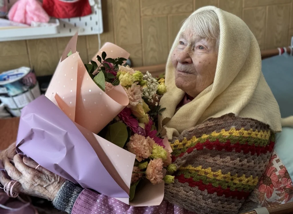 Labai sena moteris su megzta margaspalve skara ir gelsva skarele rankose laiko didelę pastelinio atspalvio gėlių puokštę.