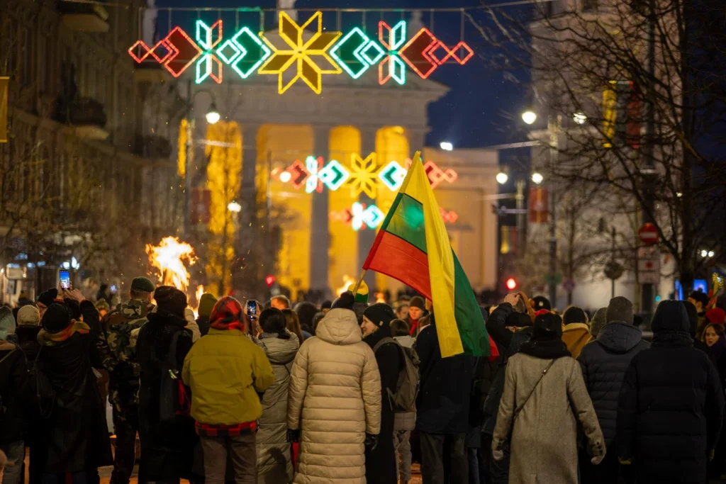Minios žmonių naktiniame Vilniuje prie laužo, plevėsuoja Lietuvos vėliava ir šviečia šventinės tautinės dekoracijos.