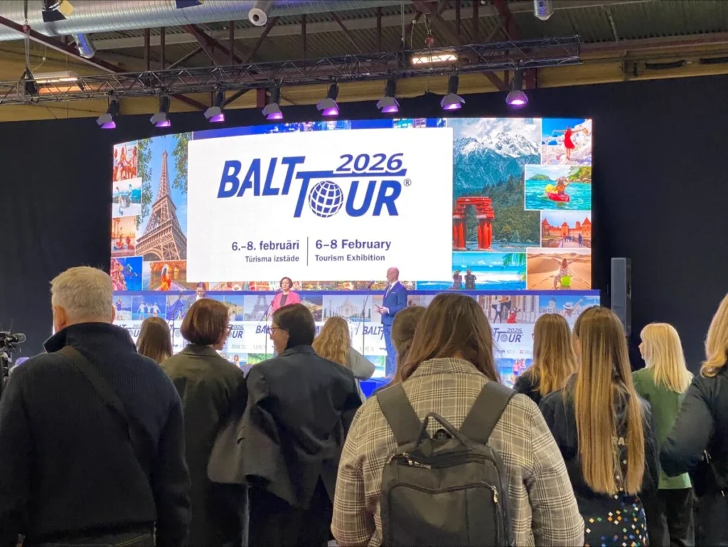 Didžiulė auditorija stovi konferencijų salėje, žiūrėdama į ryškų ekraną, skelbiantį "BALTTOUR 2026" datą.