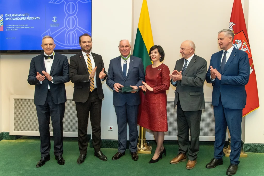 Grupė asmenų ploja apdovanojimą gavusiam vyrui oficialios ceremonijos metu.