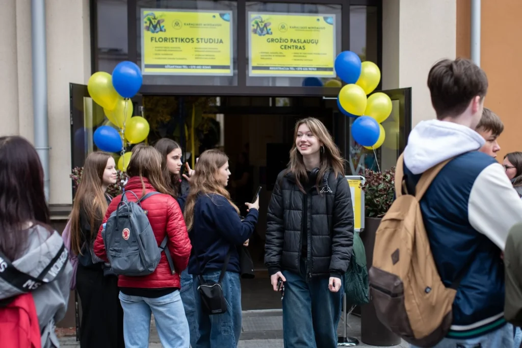 Jaunuoliai stovi prie Karaliaus Mindaugo profesinio mokymo centro durų, papuoštų mėlynais ir geltonais balionais.