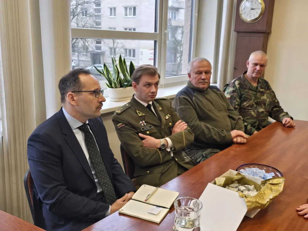 Keturi vyrai, vilkintys civiliais drabužiais ir karinėmis uniformomis, sėdi prie ilgo medinio stalo patalpoje.