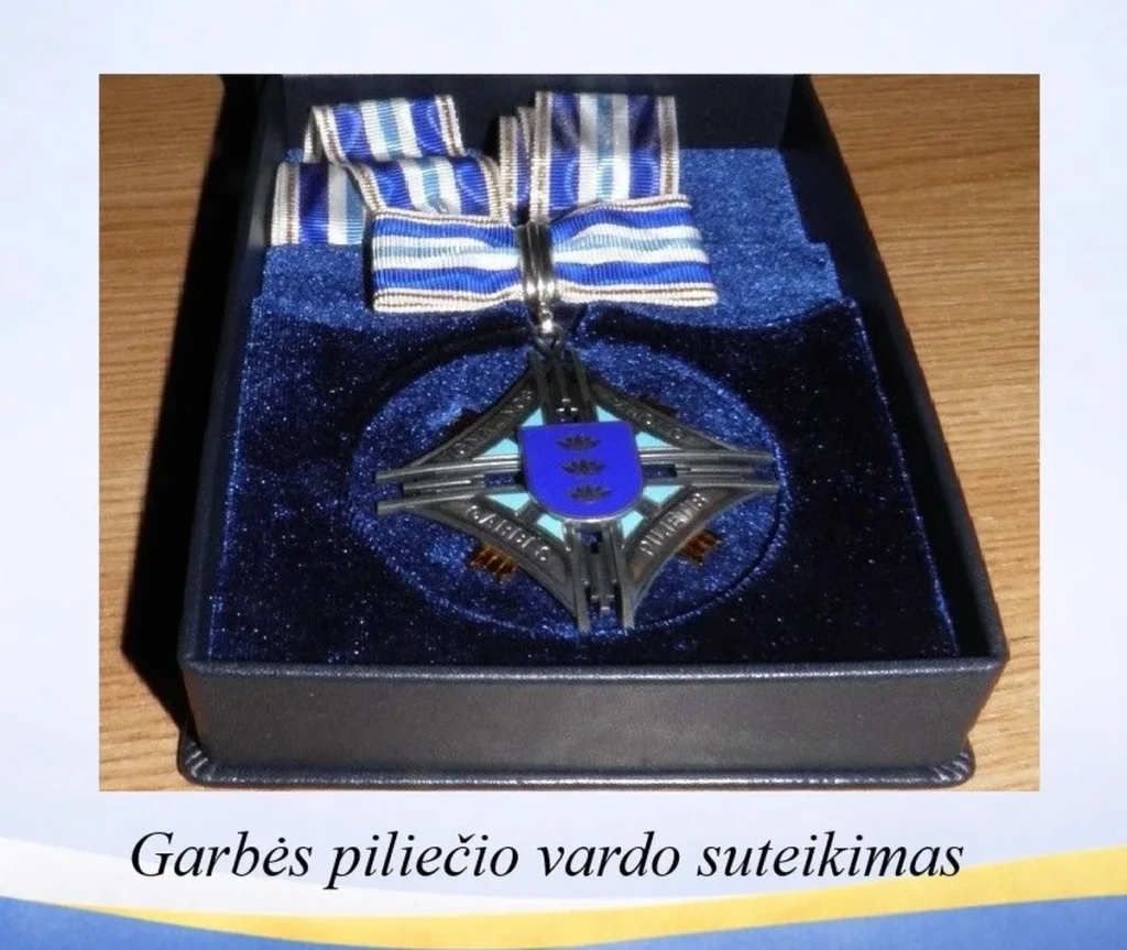 Sidabrinis garbės piliečio medalis su mėlynu herbu ir dryžuotu kaspinu tamsioje aksominėje dėžutėje.