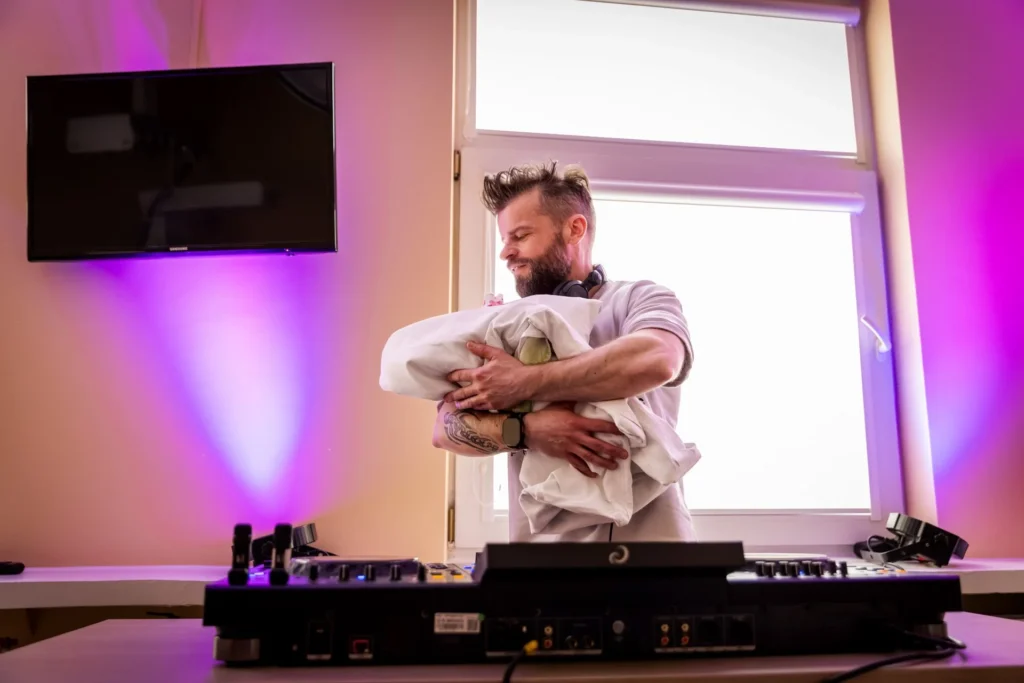 Barzdotas vyras su ausinėmis ant kaklo meiliai laiko suvystytą kūdikį, stovėdamas prie profesionalaus DJ pulto.