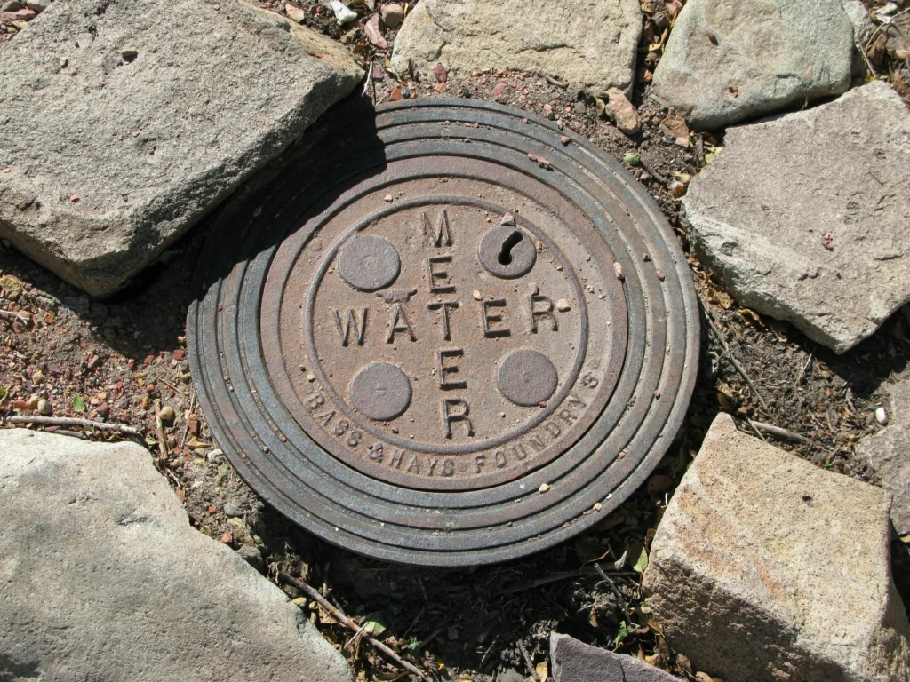 Senas ketaus vandens skaitiklio dangtis su užrašu „Water Meter“, apsuptas stambių pilkų akmenų grindinio.