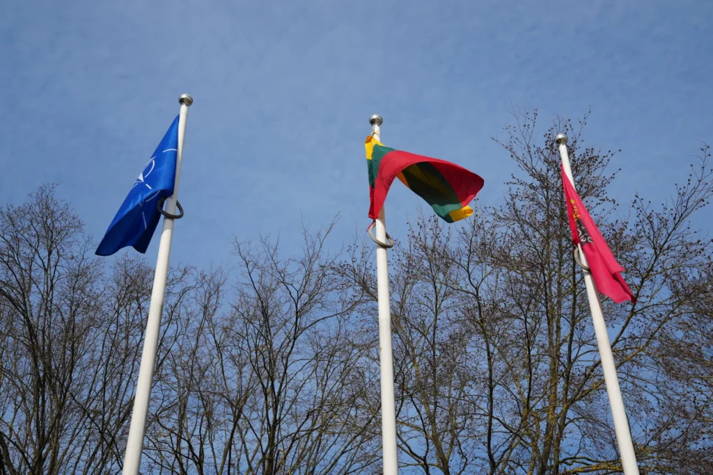 Ant baltų stiebų giedrame danguje plevėsuoja mėlyna NATO, Lietuvos trispalvė ir raudona valstybinė vėliavos.