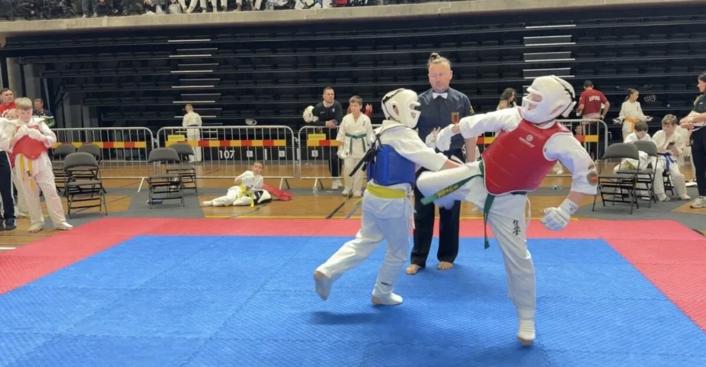 Karatė varžybose kovotojas raudona liemene atlieka aukštą smūgį koja kitam sportininkui stebint oficialiam teisėjui.