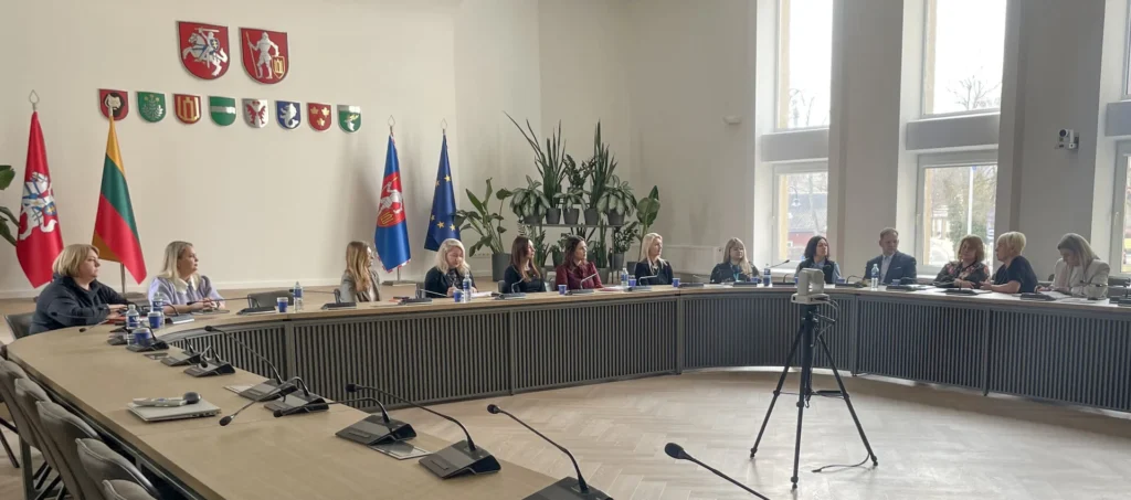 Žmonės sėdi prie pusapvalio stalo posėdžių salėje su Lietuvos vėliavomis ir herbais ant sienos.
