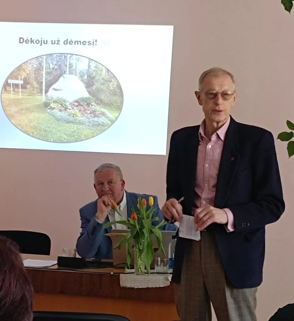 Akiniuotas vyras tamsiu švarku skaito pranešimą, fone matomas projektoriaus ekranas su užrašu „Dėkoju už dėmesį“.