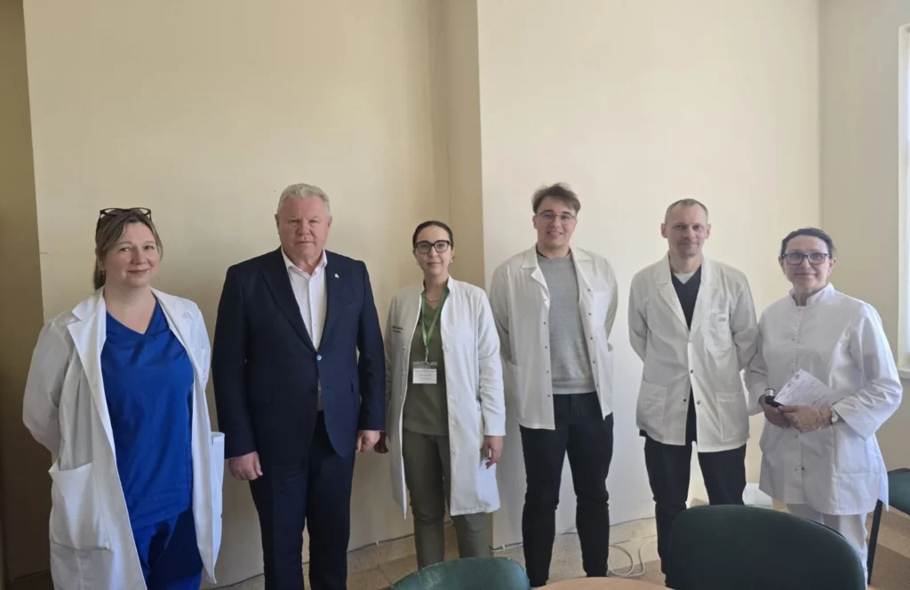Penki medicinos specialistai baltais chalatais ir kostiumuotas vyras stovi eilėje prie šviesios sienos.