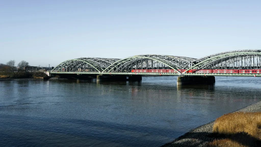 Raudonas traukinys važiuoja per masyvų žalią metalinį arkinį tiltą virš plačios upės giedrą dieną.