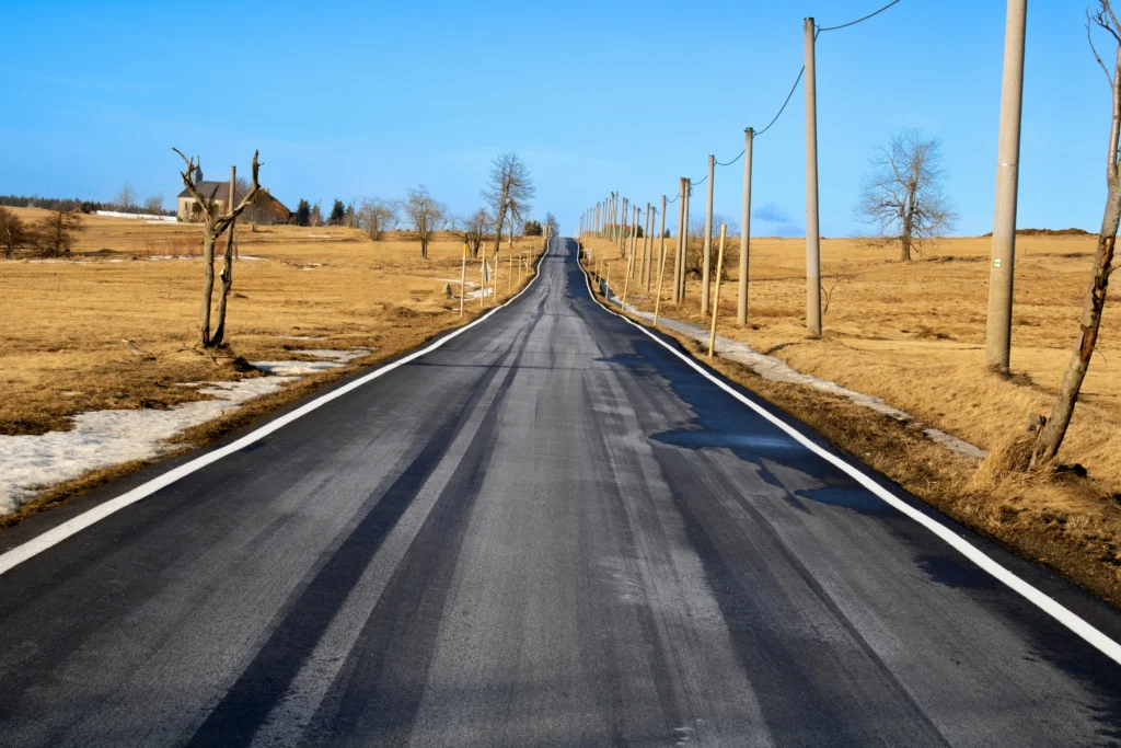 Ilgas asfaltuotas kelias driekiasi per sausus gelsvus laukus link horizonto po giedru mėlynu dangumi.