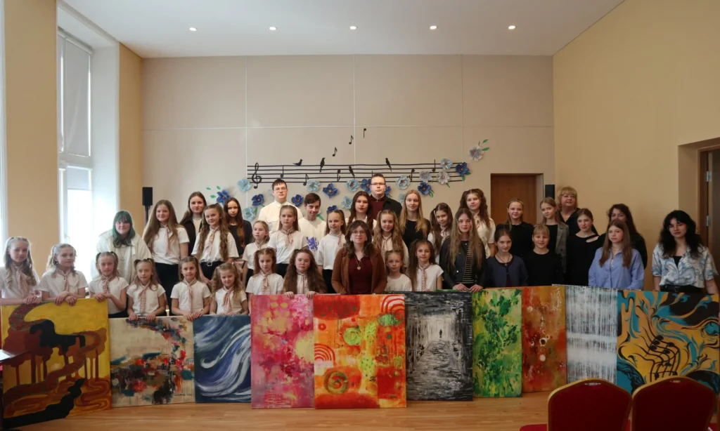 Grupė mokinių ir mokytojų stovi už ryškių abstrakčių drobių šviesioje salėje su muzikinėmis dekoracijomis.