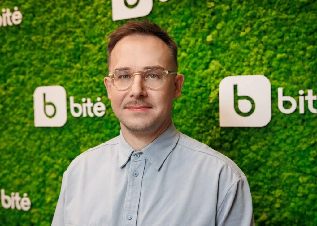 Vyras su akiniais prie žalios sienos su „Bitės“ logotipais.