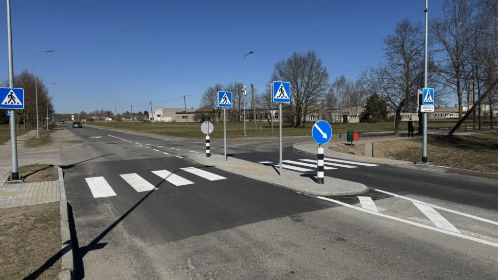 Pėsčiųjų perėja su saugumo sala, kelio ženklais ir horizontaliuoju žymėjimu Girelės gatvėje, giedrą dieną.