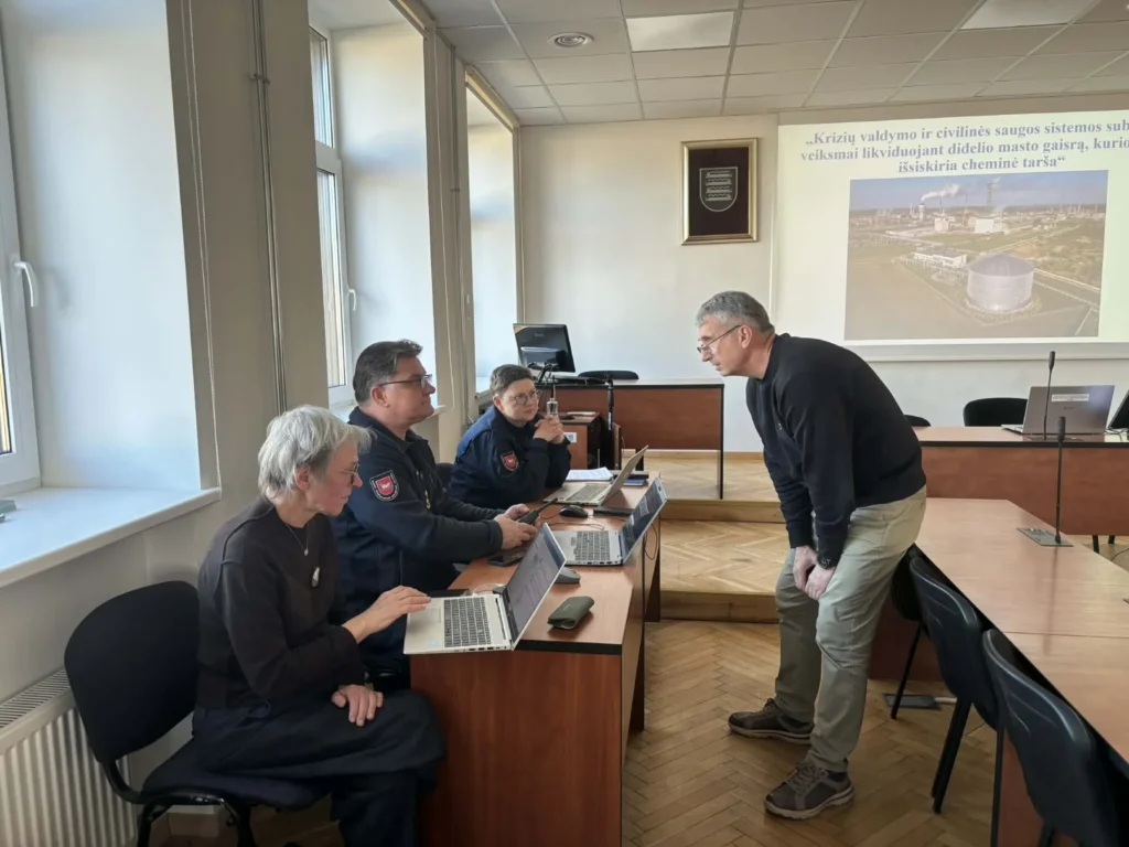 Keli asmenys ir uniformuoti pareigūnai prie nešiojamų kompiuterių aptaria civilinės saugos pratybas auditorijoje.