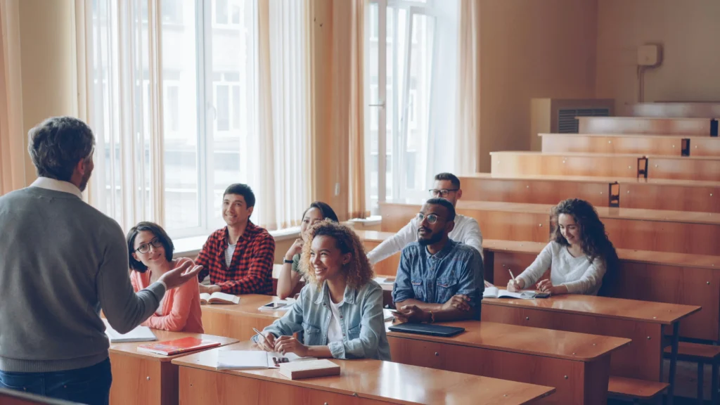 Universiteto auditorijoje sėdintys besišypsantys įvairių tautybių studentai klausosi priešais stovinčio dėstytojo dėstomos paskaitos.