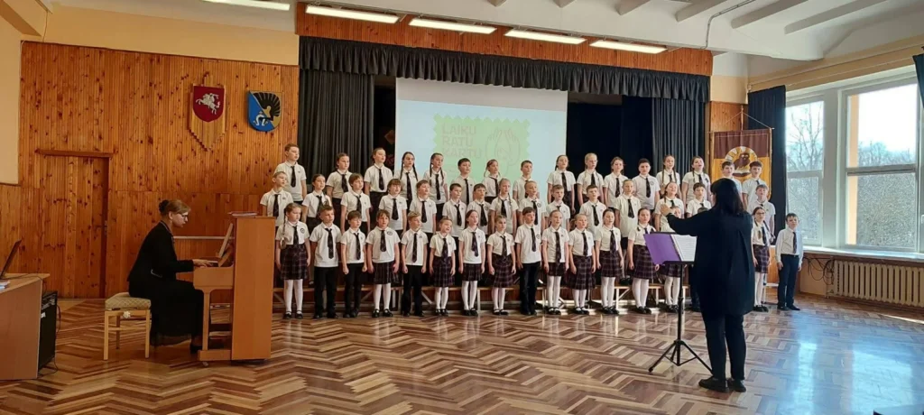 Uniformuotas vaikų choras dainuoja ant scenos, akompanuojant pianinu ir vadovaujant dirigentei jaukioje salėje.