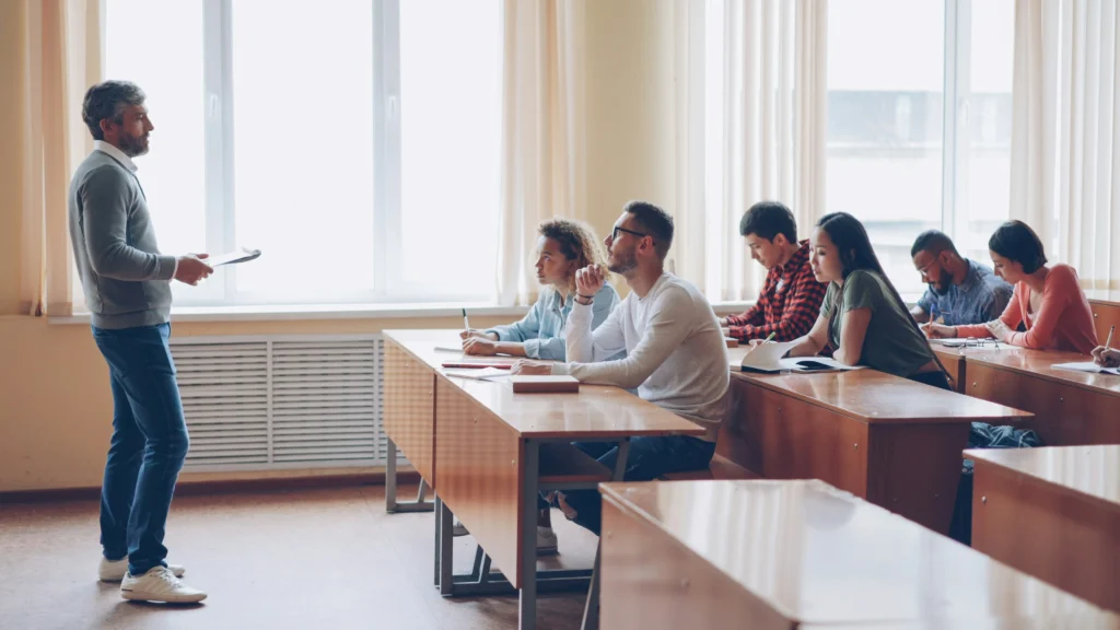 Vidutinio amžiaus dėstytojas stovi priešais grupę studentų, kurie sėdi suoluose ir klausosi paskaitos.